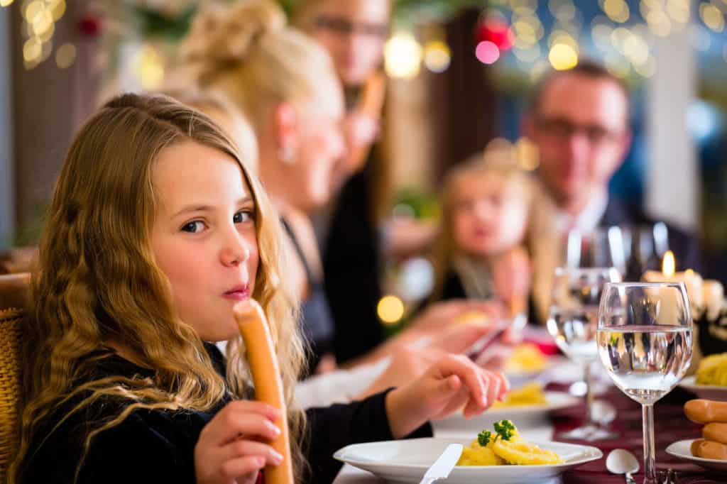 Ein Mädchen sitzt mit seiner Familie an Weihnachten am Esstisch und ist ein Wiener Würstchen. Auf ihrem Teller liegt Kartoffelsalat. Leckeres Weihnachtsessen.