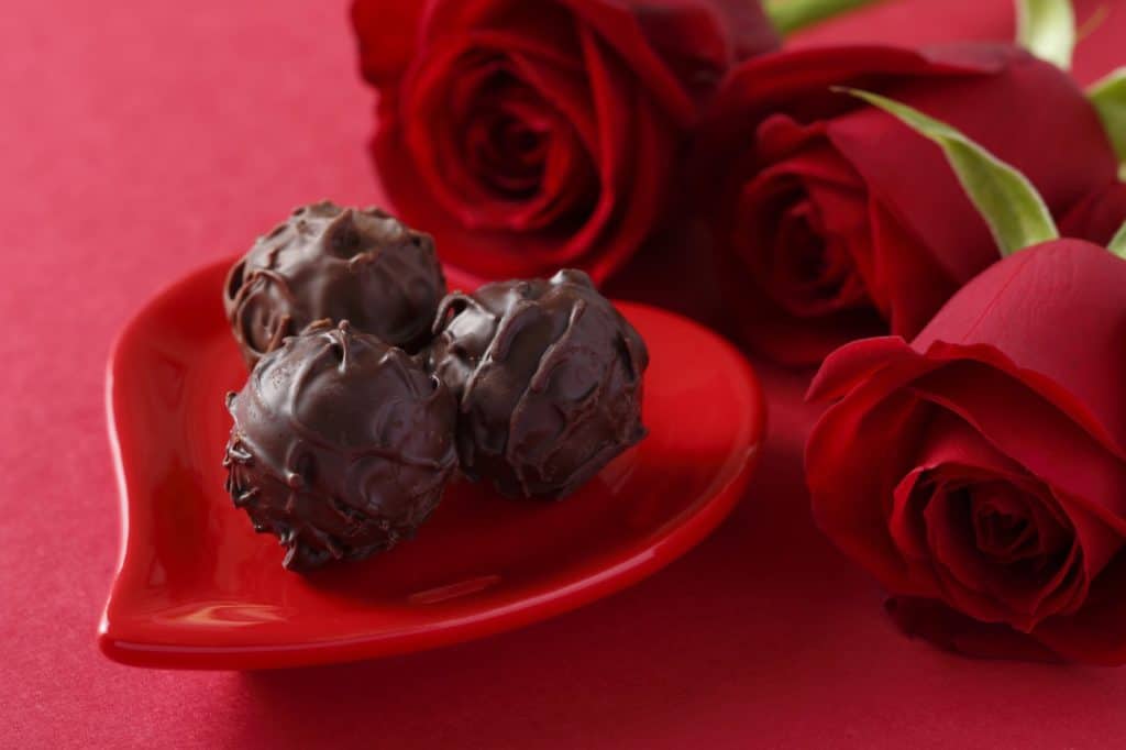 Eine kleine rote Herzschale mit Schokoladenkugeln und einem Strauß roter Rosen