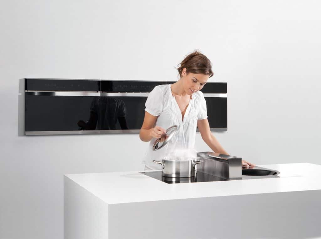 Wohnküche: Frau hebt den Glasdeckel eines Kochtopfes. In dem Kochfeld ist der Dunstabzug integriert.