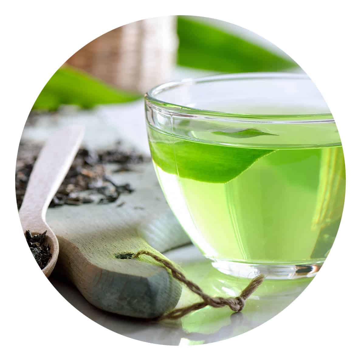Eine Tasse aus Glas gefüllt mit Grünem Tee. Naben ein Holzbrett mit losem Tee.