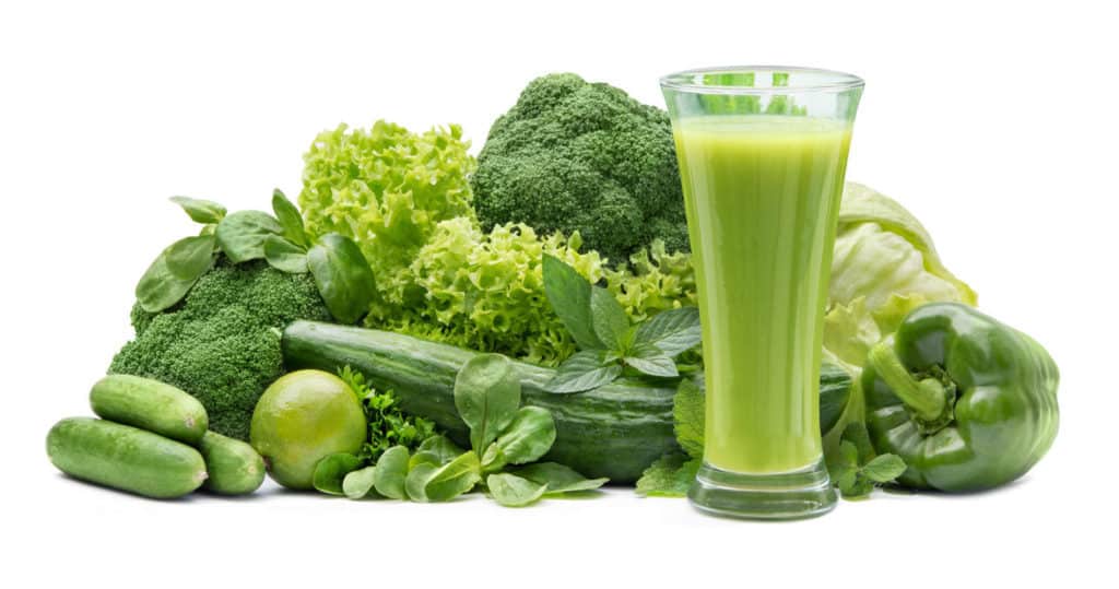 Ein grüner Smoothie im Glas vor seinen Grundzutaten wie Brokkoli, Paprika, Gurke, Salaten und Kräutern