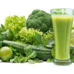 Ein grüner Smoothie im Glas vor seinen Grundzutaten wie Brokkoli, Paprika, Gurke, Salaten und Kräutern