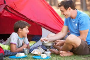 Vater und Sohn im Campingurlaub sitzen vorm Zelt und kochen mit einem Gaskocher.