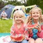 Zwei Mädchen im Campingurlaub essen Melone. Im Hintergrund ihre Eltern und das Zelt.