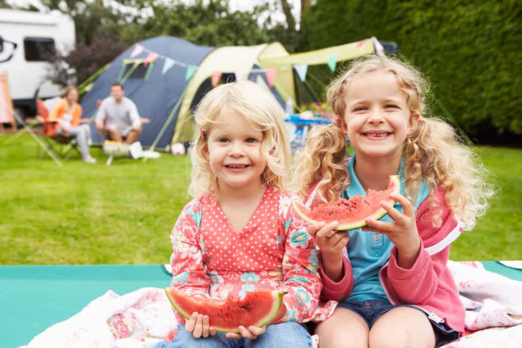 Zwei Mädchen im Campingurlaub essen Melone. Im Hintergrund ihre Eltern und das Zelt.