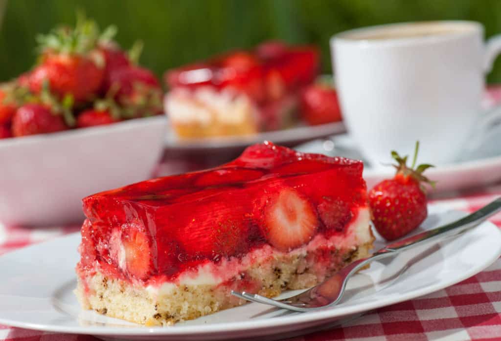 Ein Stück Erdbeertorte, im Hintergrund eine Schüssel mit frischen Erdbeeren und eine weiße Tasse – die Erdbeersaison hat begonnen.
