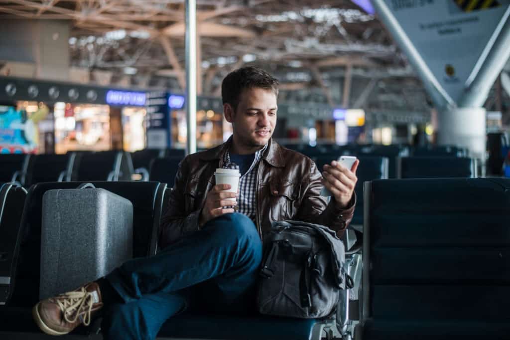 Junger Mann sitzt lächelnd auf den Stühlen des Airports, schaut auf sein Handy und hält einen Becher Kaffee – Reisen mit kleinem Budget