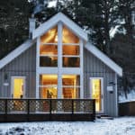 Graues Haus mit weißen Fenstern in Holzbauweise zwischen Bäumen im Schnee.