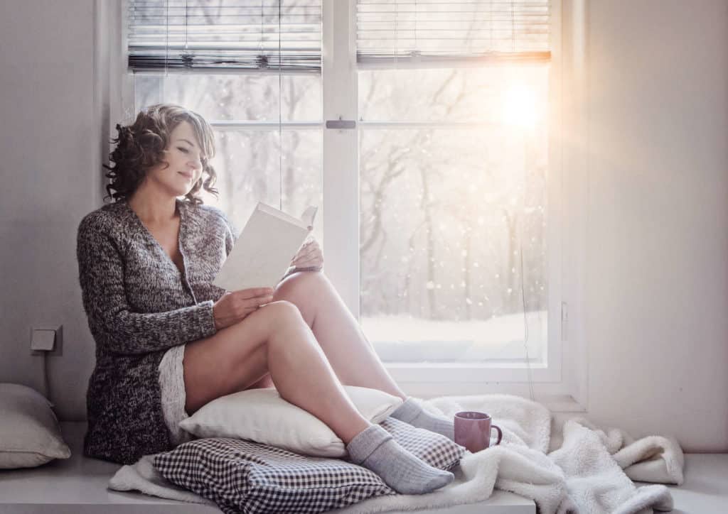 Frau sitzt in Wolljacke und Wollsocken mit nackten Beinen am Fenster bei einer Tasse Tee und liest, während die Wintersonne ins Haus scheint.