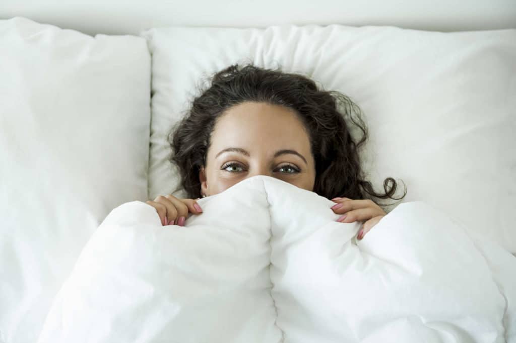 Brünette Frau liegt sich im weiß bezogenen Bett und hat sich die Bettdecke bis über die Nase hochgezogen.