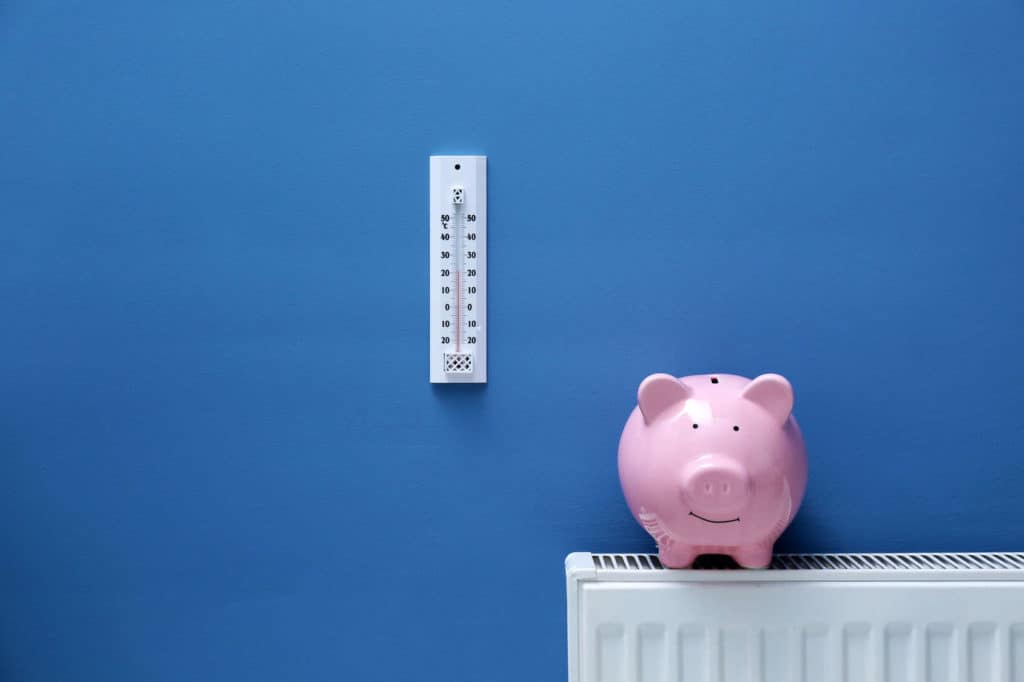 Ein rosa Sparschwein steht auf einer Heizung vor blauem Hintergrund, an der Wand hängt ein Thermostat.
