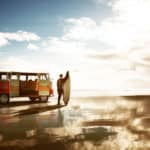Sanfter Tourismus: Surfer mit Surfbrett vor einem VW-Bus schaut in die Wellen und die Sonne.