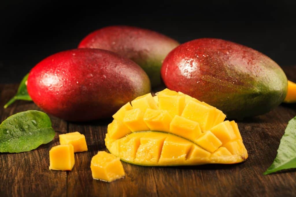 Lebensmittel-Lifehacks, Nummer 1: Drei Mangos und eine halbe, aufgeschnittene Mango liegen auf einem dunklen Holztisch.