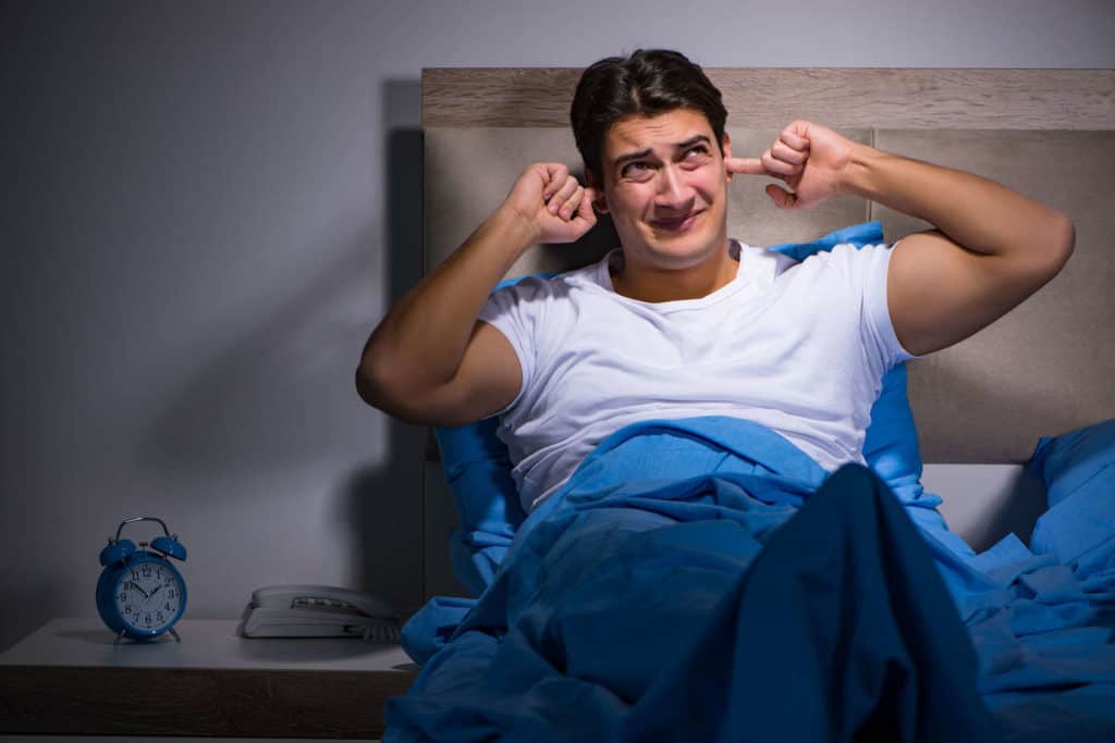 Lärm im Schlafzimmer: Ein Mann sitzt nachts im Bett und hält sich die Ohren zu.