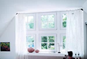 Nach dem Fensteraustausch ist vor der Pflege: Mehrere kleinere weiße Kunststoff-Fenster, die in den Garten hinaus führen, umrahmt von weißen Gardienen.