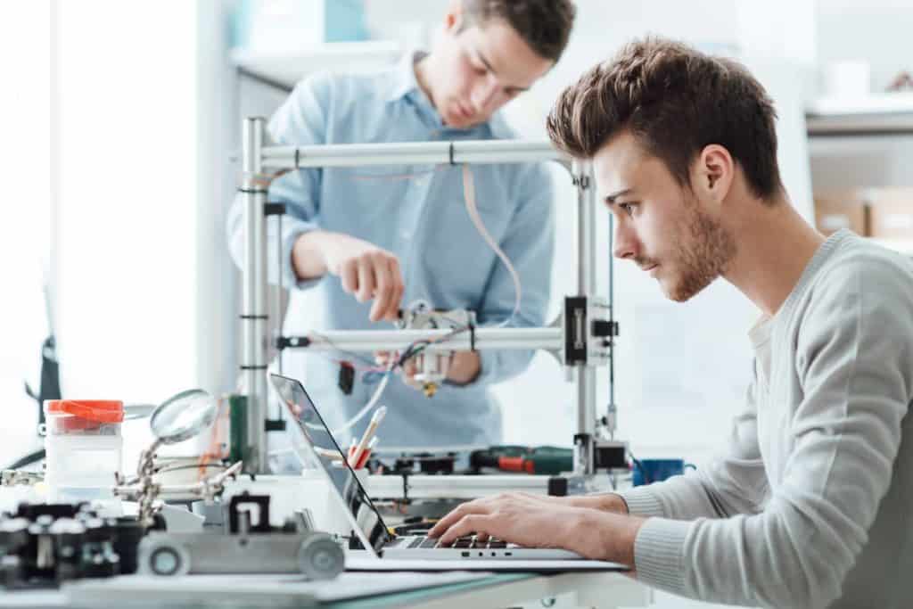Moderne Technik: Junger Mann sitzt am Laptop und programmiert einen Computer, im Hintergrund baut ein Mann einen Rechner zusammen.