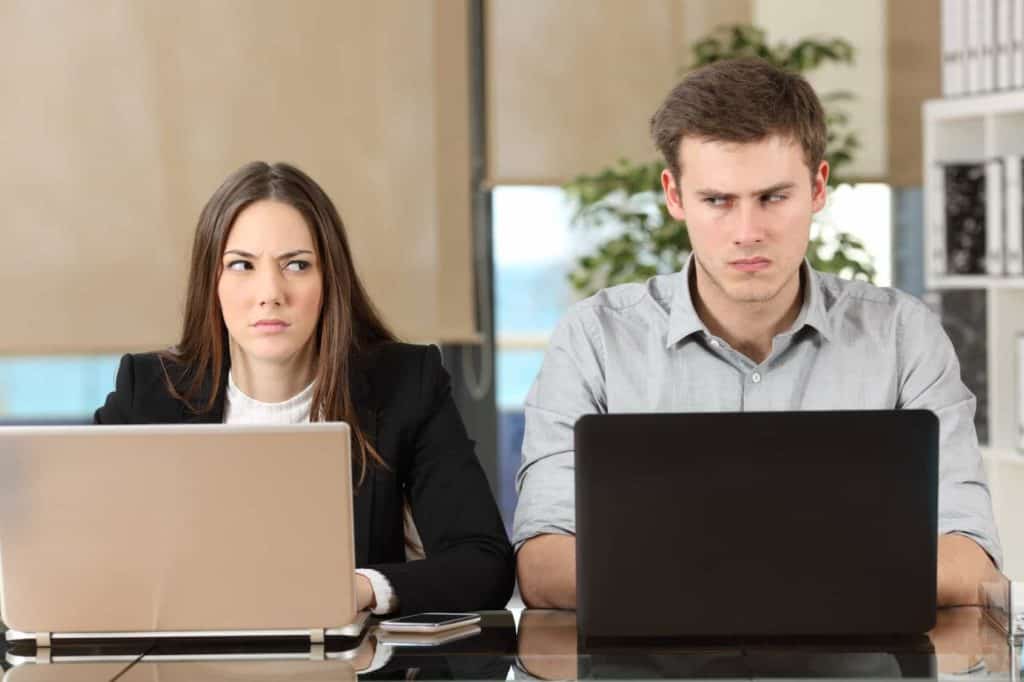 Mann und Frau sitzen vor ihren Laptops und werfen sich böse Blicke zu (nervige Kollegen)
