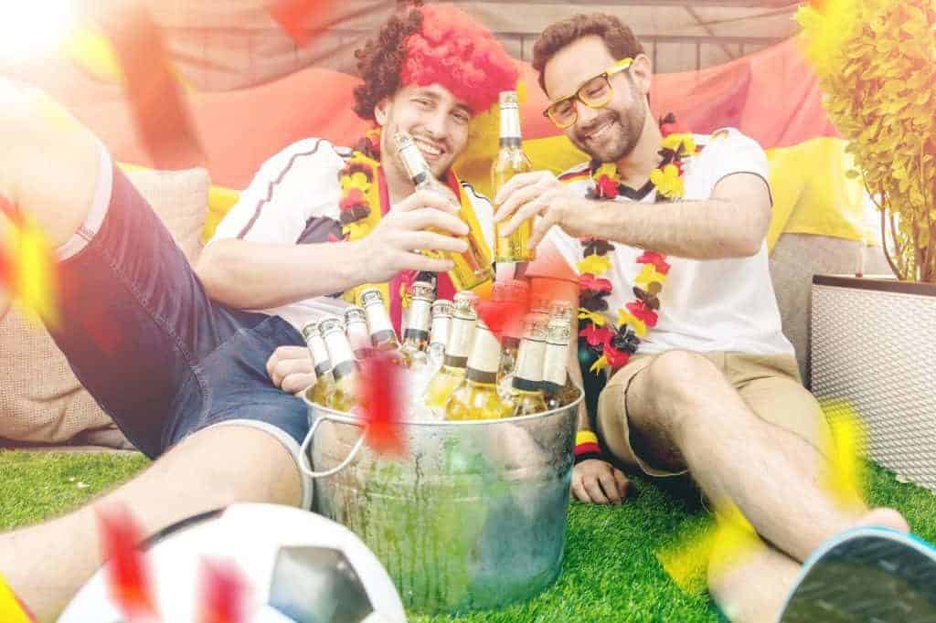 Zwei Männer in Fanbekleidung sitzen auf dem Rasen vor einem Eimer mit gekühlten Bierflaschen (WM-Party im Garten)