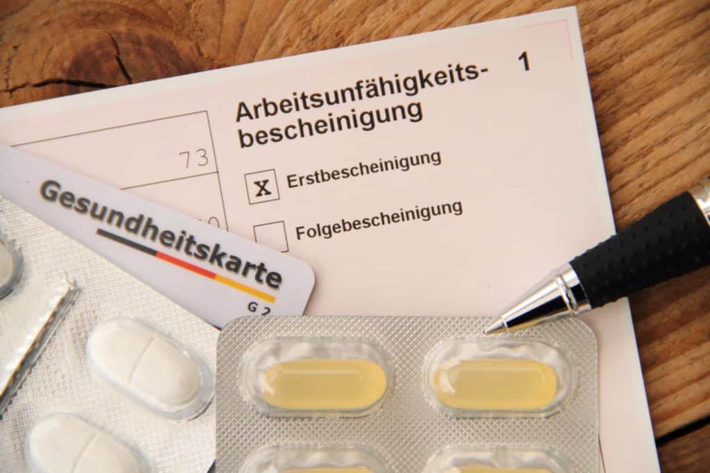 Eine Arbeitsunfähigkeitsbescheinigung für den Arbeitgeber sowie eine Versichertenkarte und Tabletten liegen neben einem Kugelschreiber auf einem Schreibtisch aus Holz