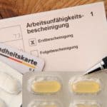 Eine Arbeitsunfähigkeitsbescheinigung für den Arbeitgeber sowie eine Versichertenkarte und Tabletten liegen neben einem Kugelschreiber auf einem Schreibtisch aus Holz