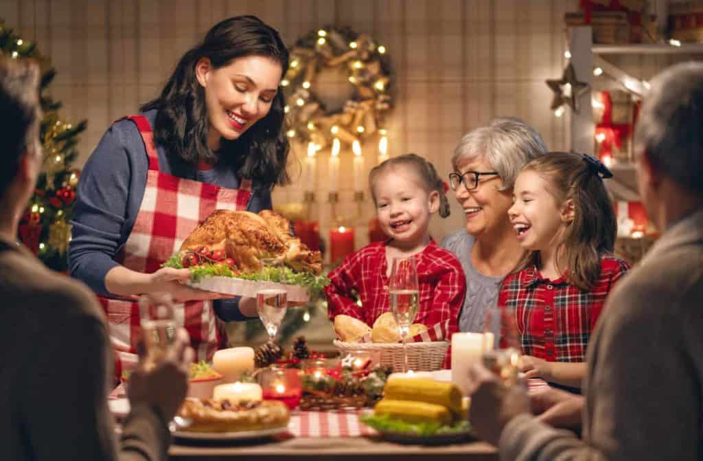 Eine Familie sitzt am weihnachtlich gedeckten Tisch, lachen gemeinsam und freuen sich über das Weihnachtsessen, welches von einer Frau serviert wird. (Weihnachten in anderen Ländern)