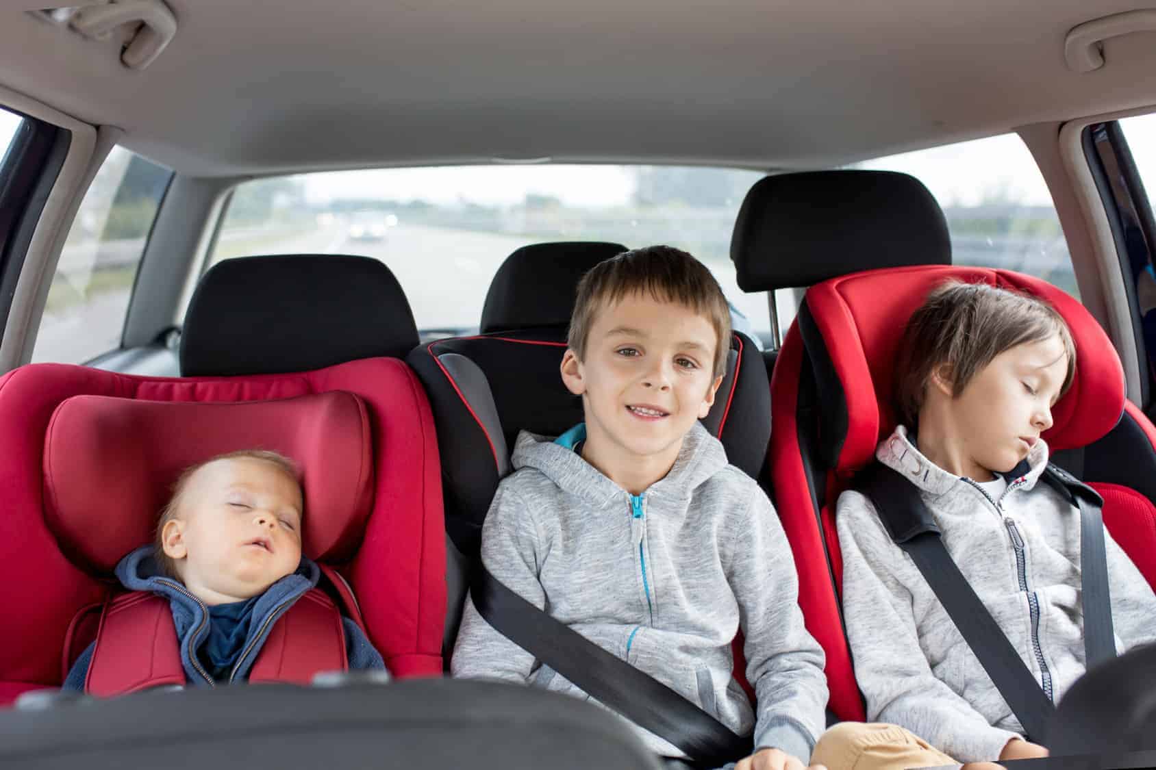 Drei Kinder sitzen auf der Rückbank eines PKWs. Während zwei schlafen, schaut einer aus dem Fenster. (Autofahren mit Kindern)