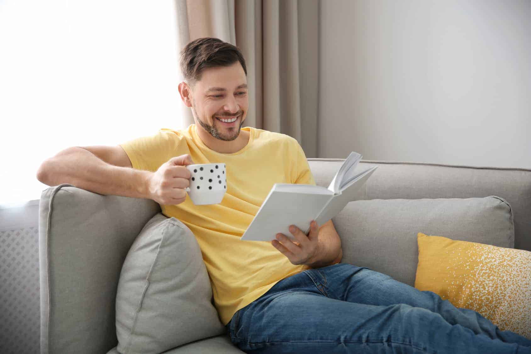 Ein Mann sitzt auf einer Couch, trinkt Tee und liest ein Buch. (Urlaub zuhause)