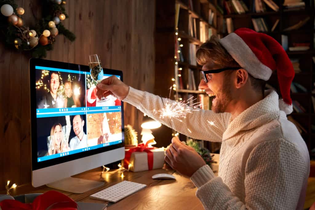 Virtuelle Weihnachtsfeier: ein lachender Mann trägt eine rote Weihnachtsmütze, hält eine gezündete Wunderkerze und ein gefülltes Sektglas in den Händen und stoßt mit seinen Kollegen in einem Video-Call an.