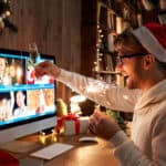 Virtuelle Weihnachtsfeier: ein lachender Mann trägt eine rote Weihnachtsmütze, hält eine gezündete Wunderkerze und ein gefülltes Sektglas in den Händen und stoßt mit seinen Kollegen in einem Video-Call an.