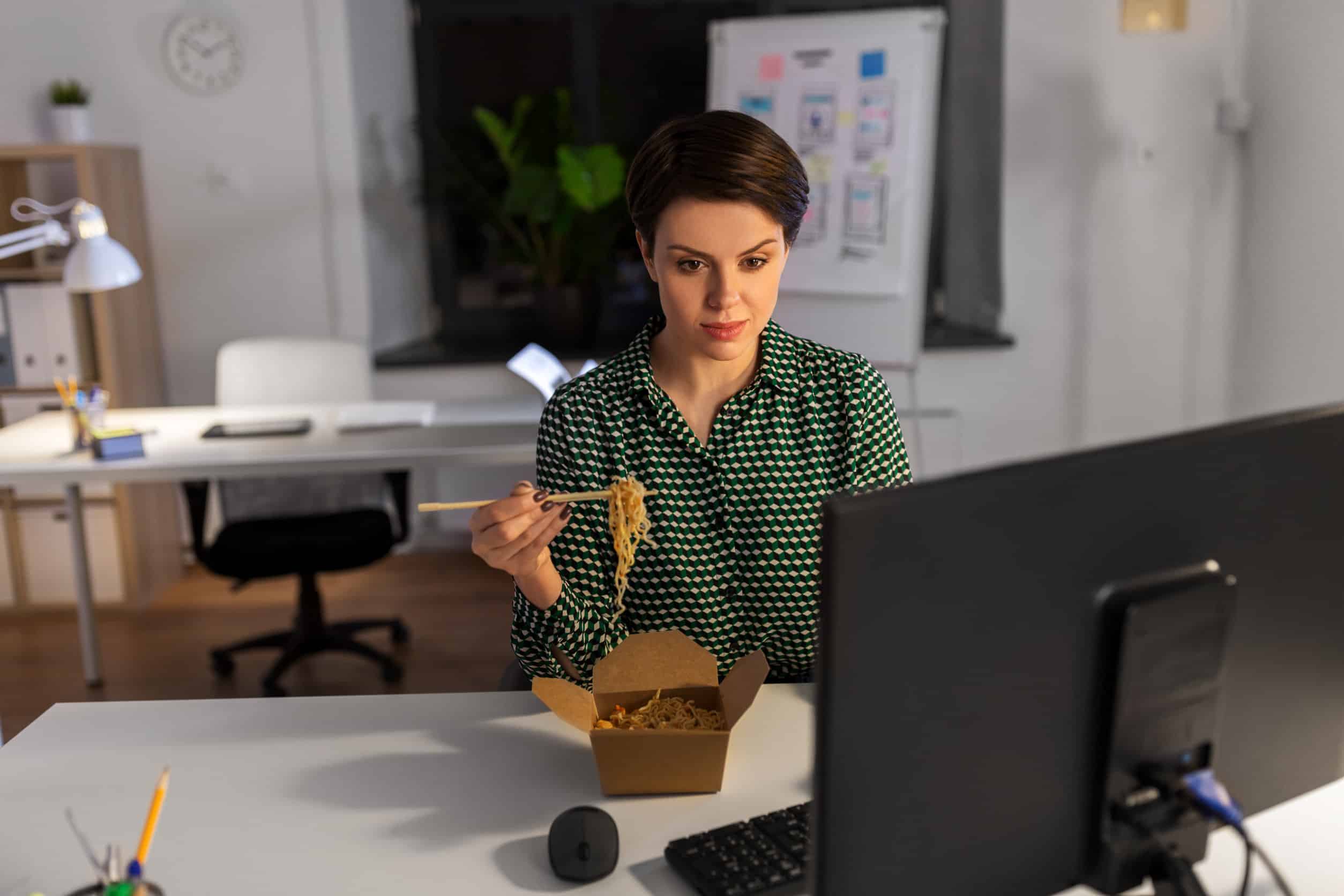 Workaholic: eine Frau sitzt vor einem Computer und isst nebenbei gebratene Nudeln aus der To-Go-Box.
