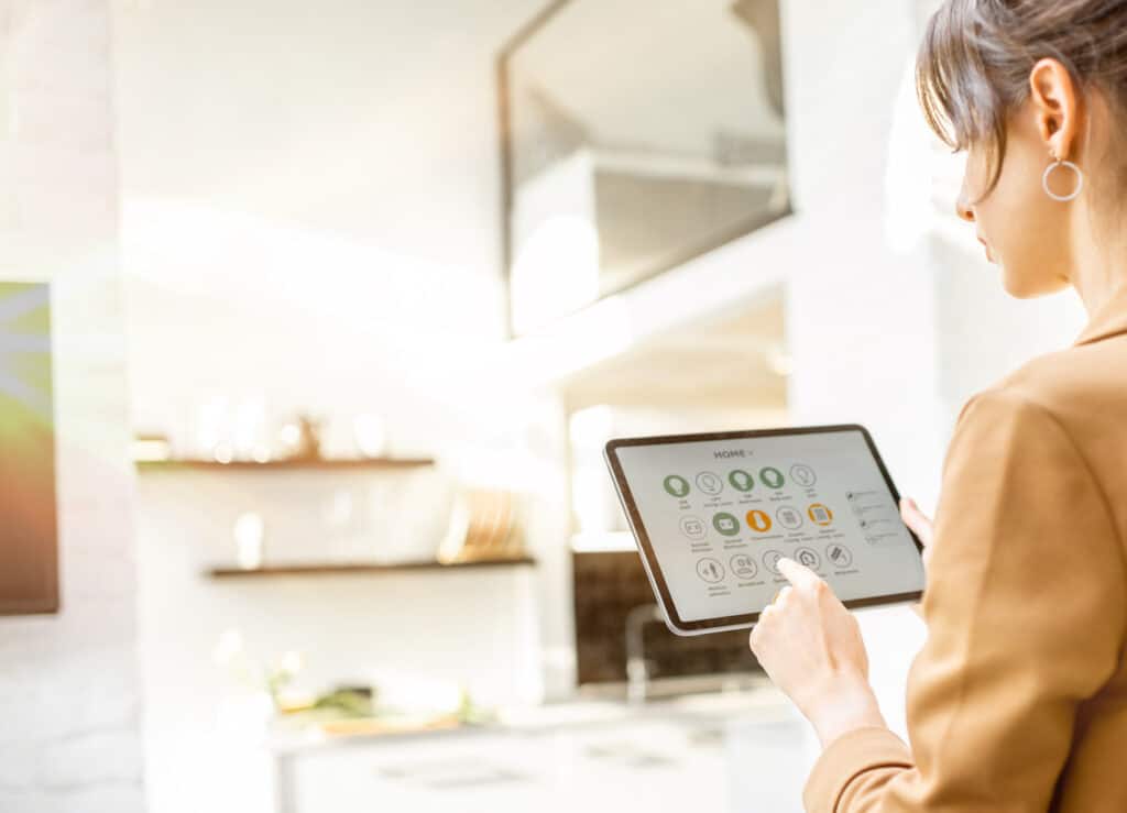 Eine weibliche Hand hält ein Tablet, auf die Steuerungs-Icons einer Smart Home-App zu sehen sind. steht unter einem Spiegel ein Paar Schuhe.