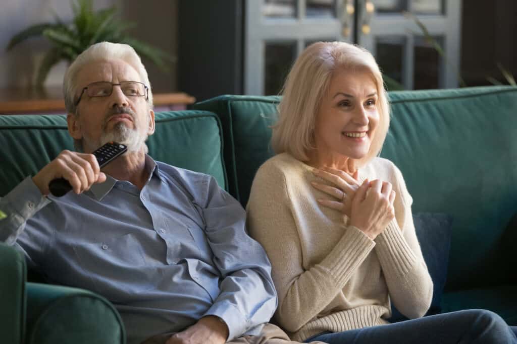 Ein älteres Ehepaar sitzt auf dem Sofa und schaut Fernsehen. Der Mann hat Schwierigkeiten die Dialoge zu verstehen.