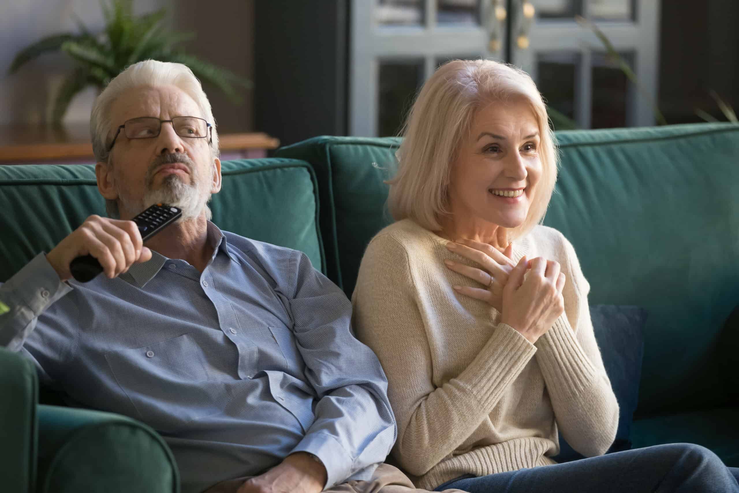 Ein älteres Ehepaar sitzt auf dem Sofa und schaut Fernsehen. Der Mann hat Schwierigkeiten die Dialoge zu hören.