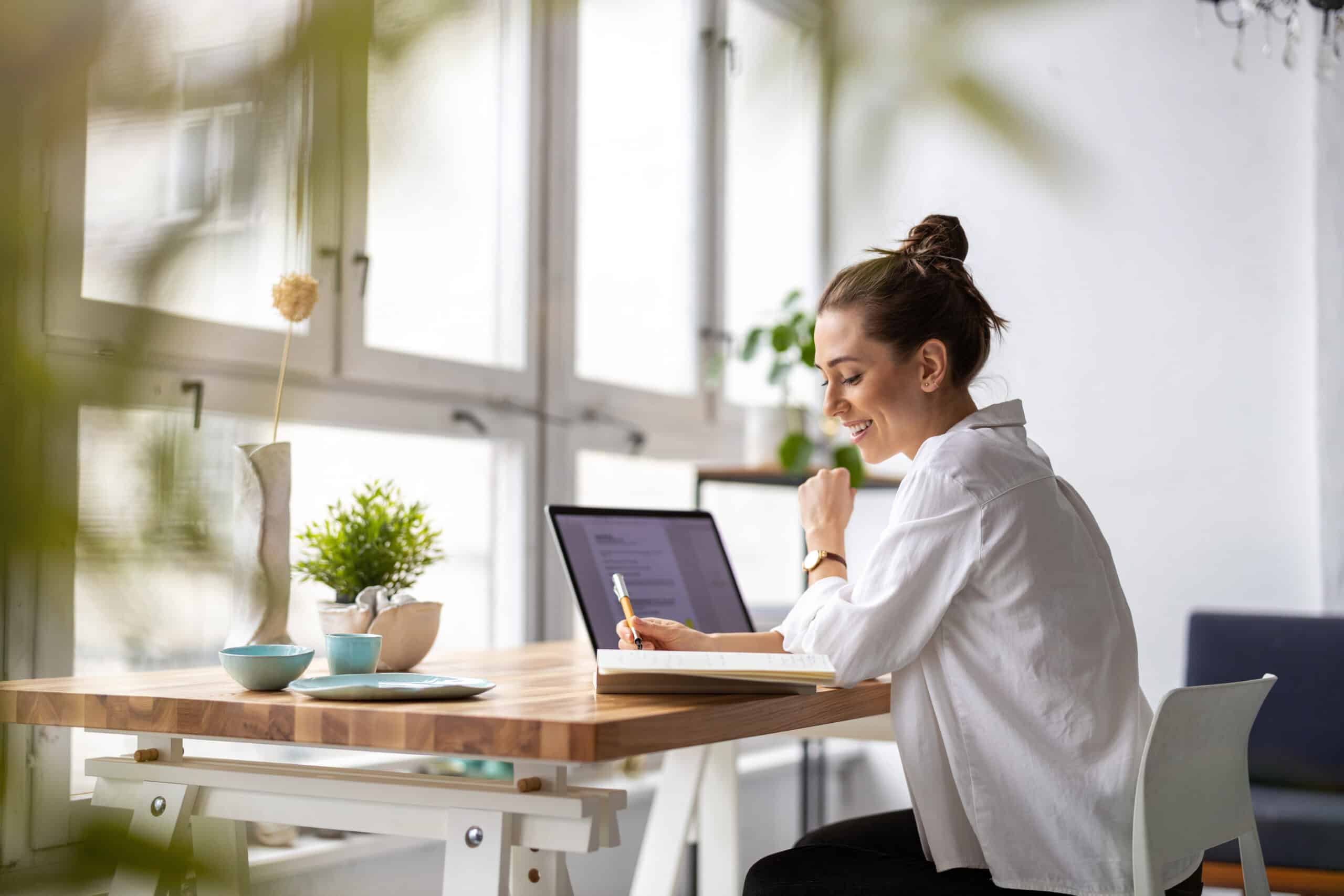 Frau sitzt an einem Schreibtisch vor vielen Fenstern und arbeitet lächenlnd an ihrem Laptop. (Workation)
