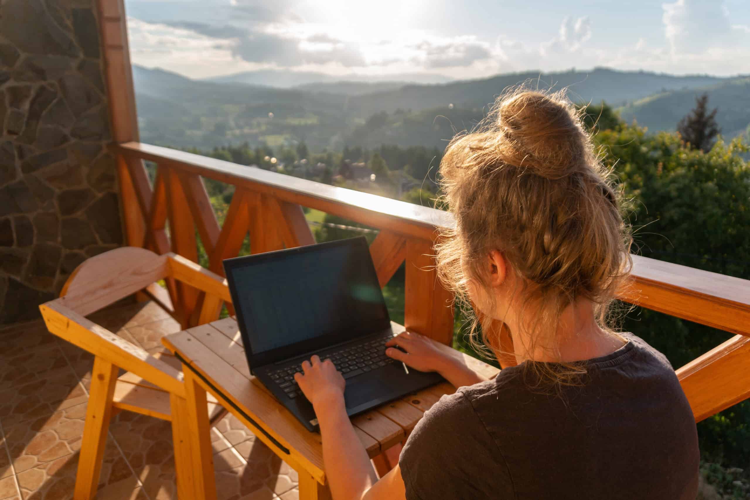 Frau arbeitet am Laptop auf dem Balkon mit einem tollen Aublick. (Workation)