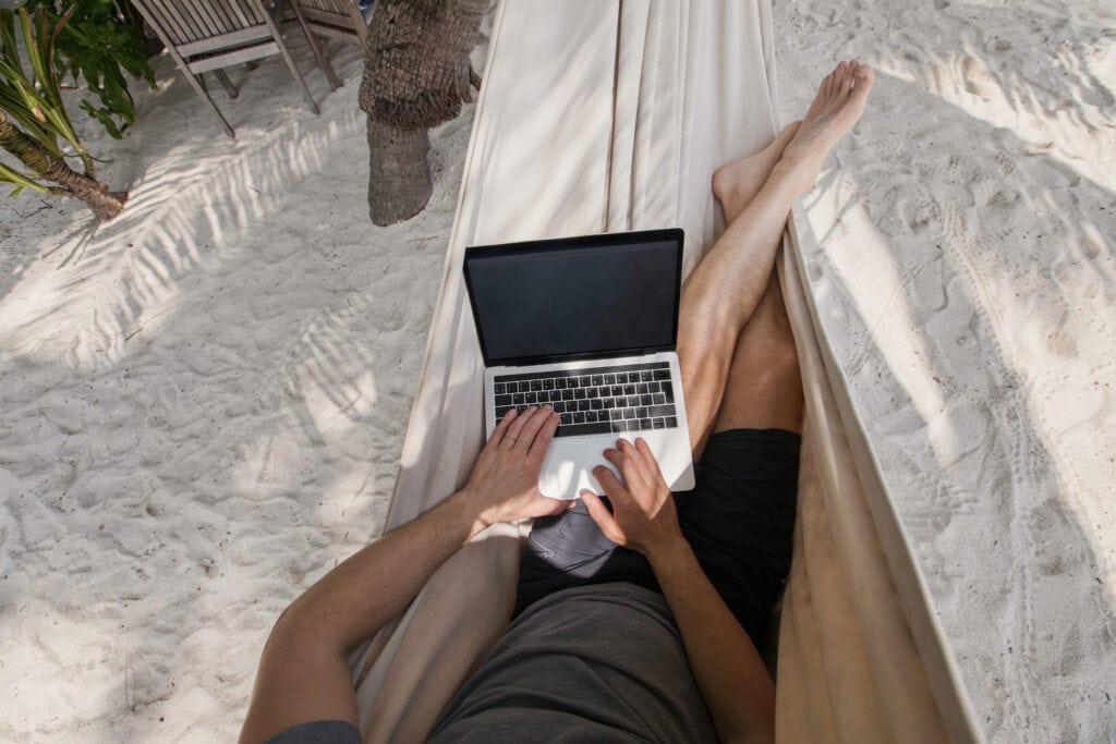 Ein Mann liegt am Strand in einer Hängematte und arbeitet mit seinem Laptop. (Workation)