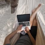 Ein Mann liegt am Strand in einer Hängematte und arbeitet mit seinem Laptop. (Workation)