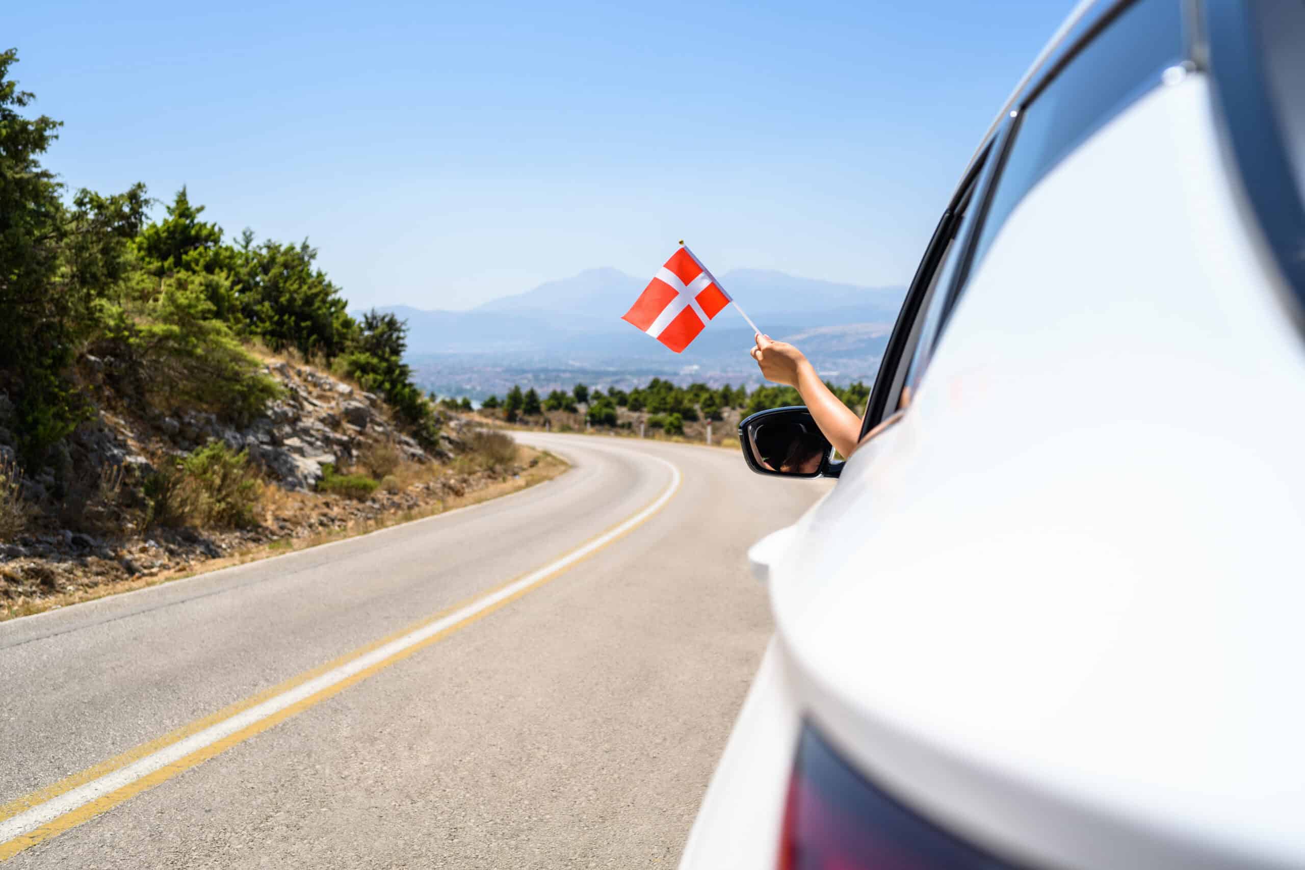 Ein Mensch hält eine Dänemarkflagge aus dem Auto auf einer Straße. (Dänemark-Urlaub)