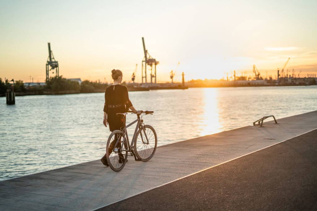 Eine Frau spaziert mit einen Fahrrad am Hamburger Hafen entlang. Die Sonne geht dabei unter.