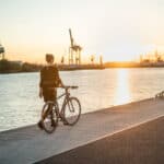 Eine Frau spaziert mit einen Fahrrad am Hamburger Hafen entlang. Die Sonne geht dabei unter.