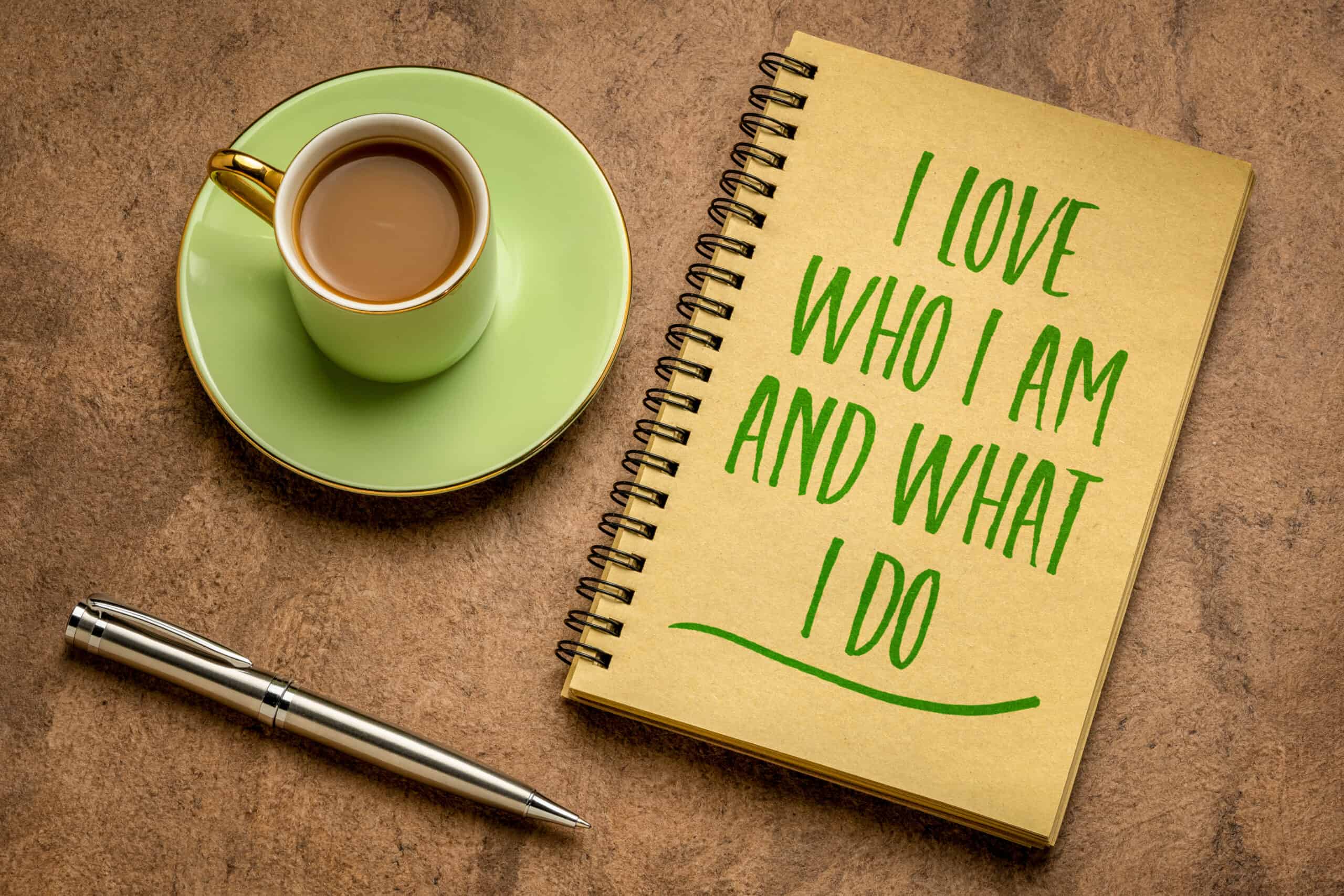 Positive Gedanken im Job: Ein Buch mit der Aufschrift "I love who I am ans what I do" mit Stift und Kaffee liegt auf einem Tisch. 