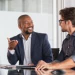 Zwei Männer sitzen an einem Arbeitstisch und unterhalten sich lächelnd.