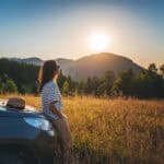 Wochendtrip in die Berge: Frau guckt sich den Sonnenuntergang bei den Bergen mit ihrem Auto an.