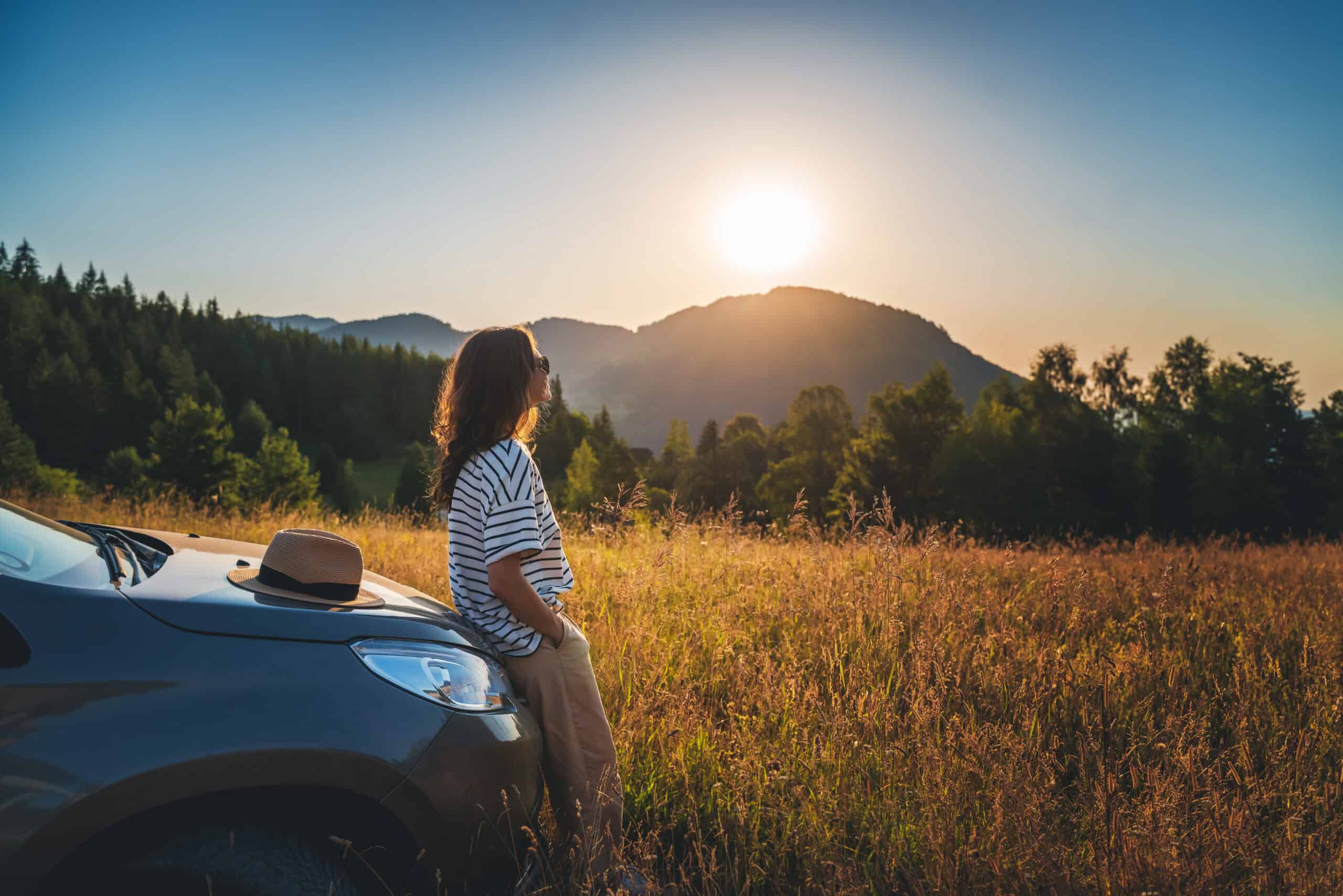 Wochendtrip in die Berge: Frau guckt sich den Sonnenuntergang bei den Bergen mit ihrem Auto an.