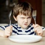 Mealprep für Einsteiger: Ein Junge zwischen 5 und 7 Jahren sitzt verärgert am Küchentisch, mit Messer und Gabel in der Hand. Vermutlich wartet er auf sein Essen.