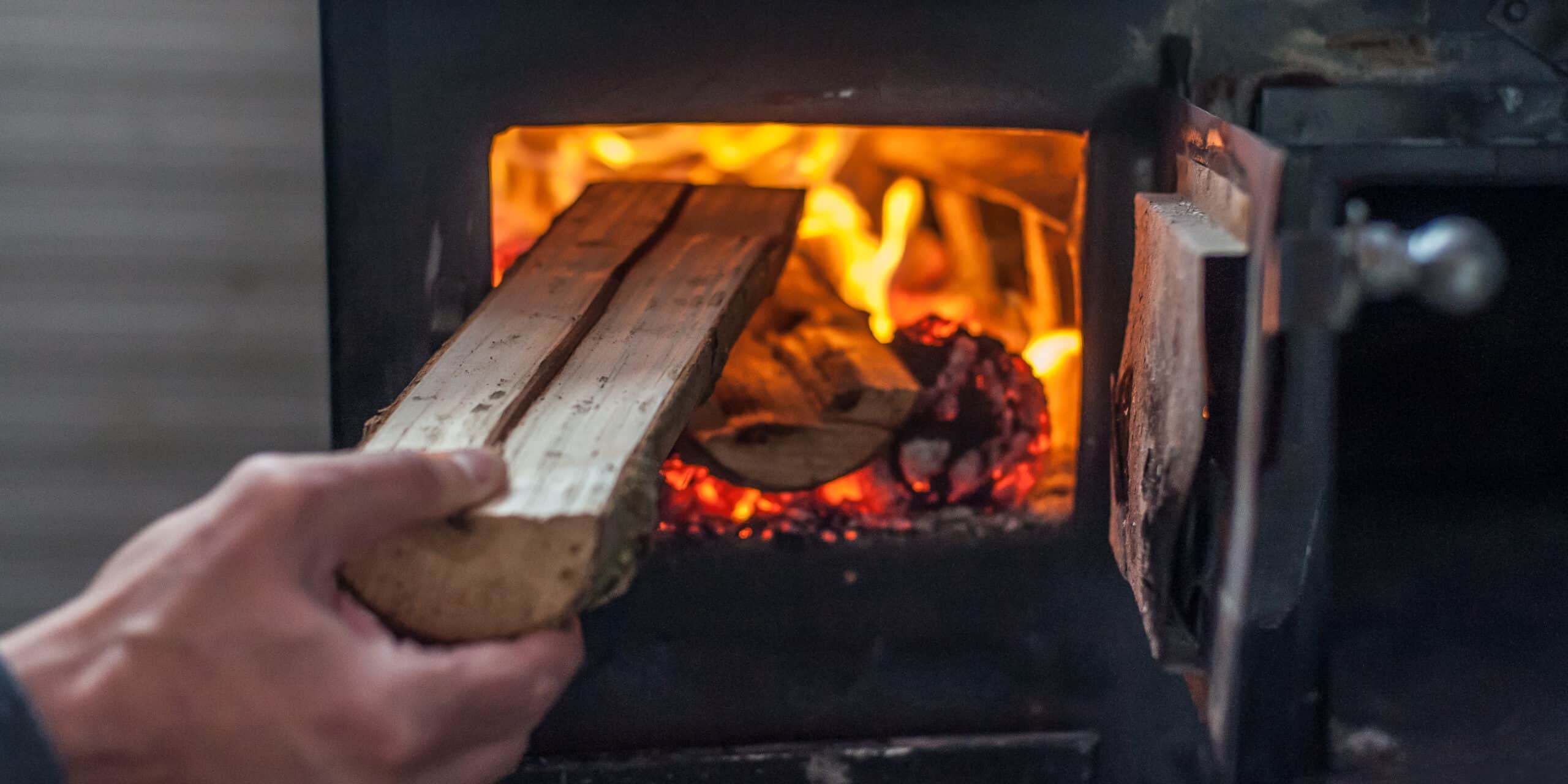 Kamine und Feuerstätten: Das sollten Hausbesitzer wissen Kamine und Feuerstätten: Eine Hand legt ein Stück Holz in den brennenden Ofen nach.