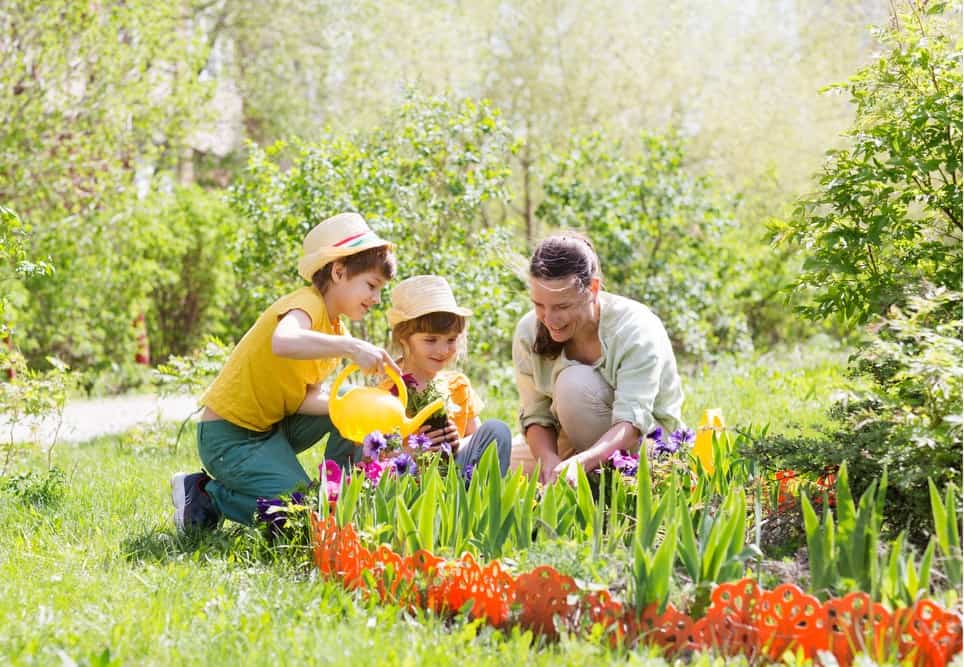 Eine Frau und zwei Kinder gärtnern gemeinsam in einem blühenden Garten und gießen Blumen.
